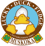 The Lucky Duck logo featuring a smiling, white duck with yellow-orange webbed feet and duck bill, with a blue background and surrounded by a ring, reading "Lucky Duck Fudge...established 1989" and beneath that it reads, "Muskoka" inbetween 2 lucky clovers